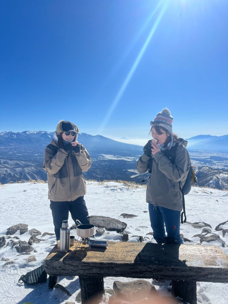 Two guests sharing a hot drink on a snowy summit with Mt. Fuji on the horizon